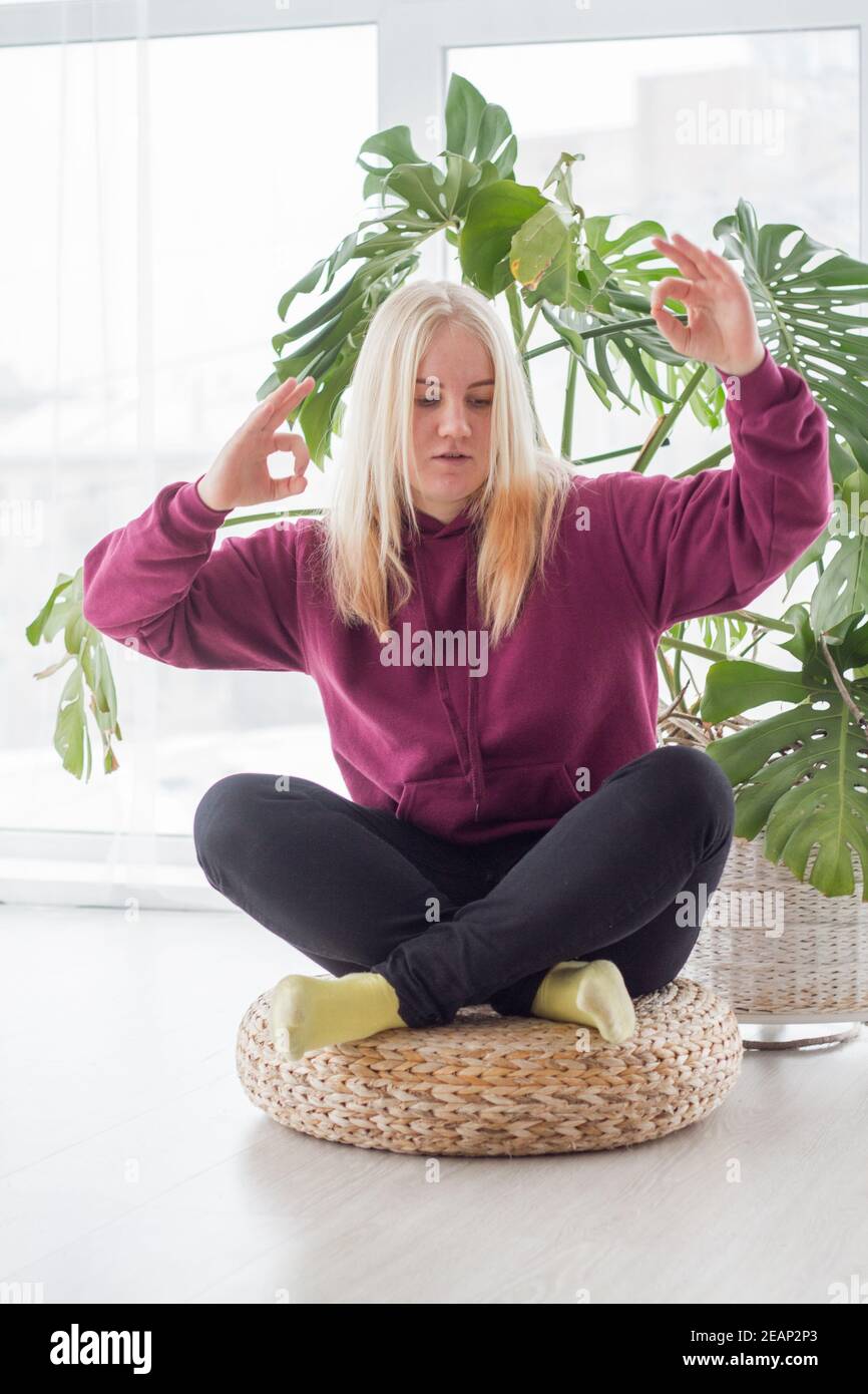 Young blonde woman meditating in lotus pose at home, practicing yoga indoors, healthy lifestyle concept Stock Photo