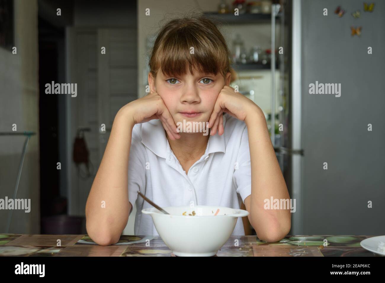 The girl is sitting at the table in the kitchen, in front of her is an empty plate, the girl looks into the frame Stock Photo