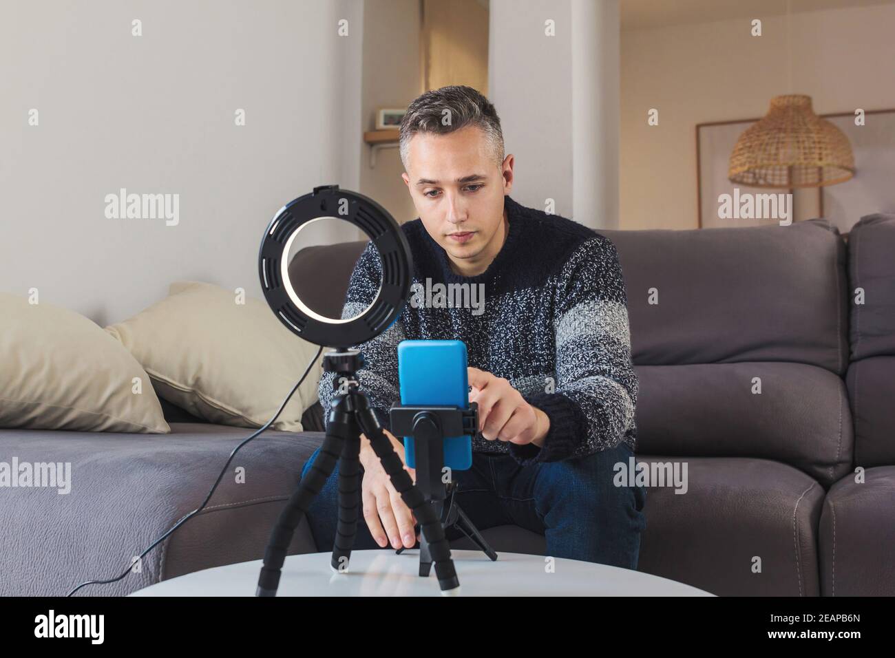 Stock photo of Young man millennial influencer sit on sofa preparing a setup with mobile phone and lighting for vlogging or video call. Social media v Stock Photo