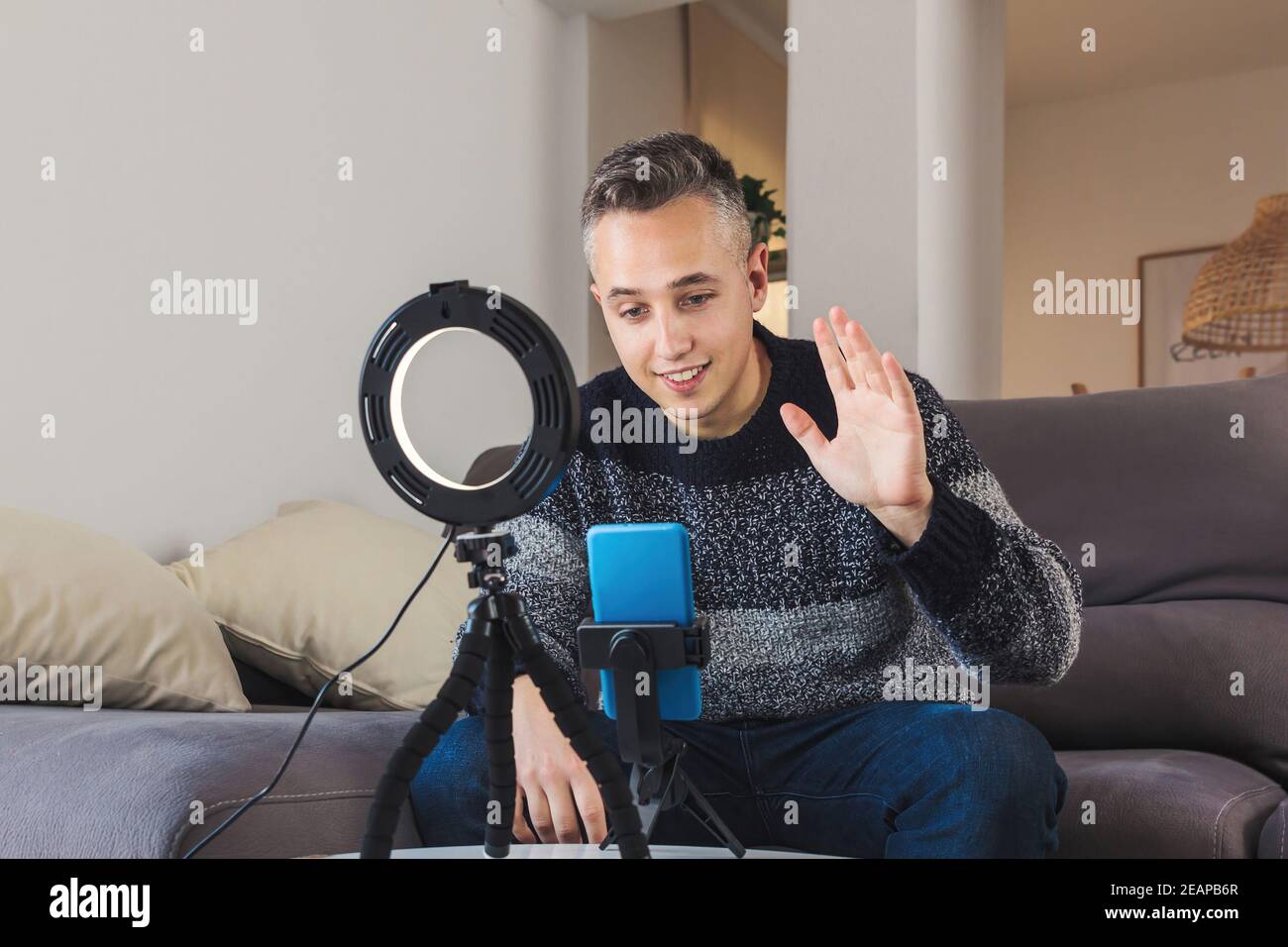 Stock photo of Young man millennial influencer sit on couch is vlogging, recording and creating online content with smartphone and lights. Social medi Stock Photo