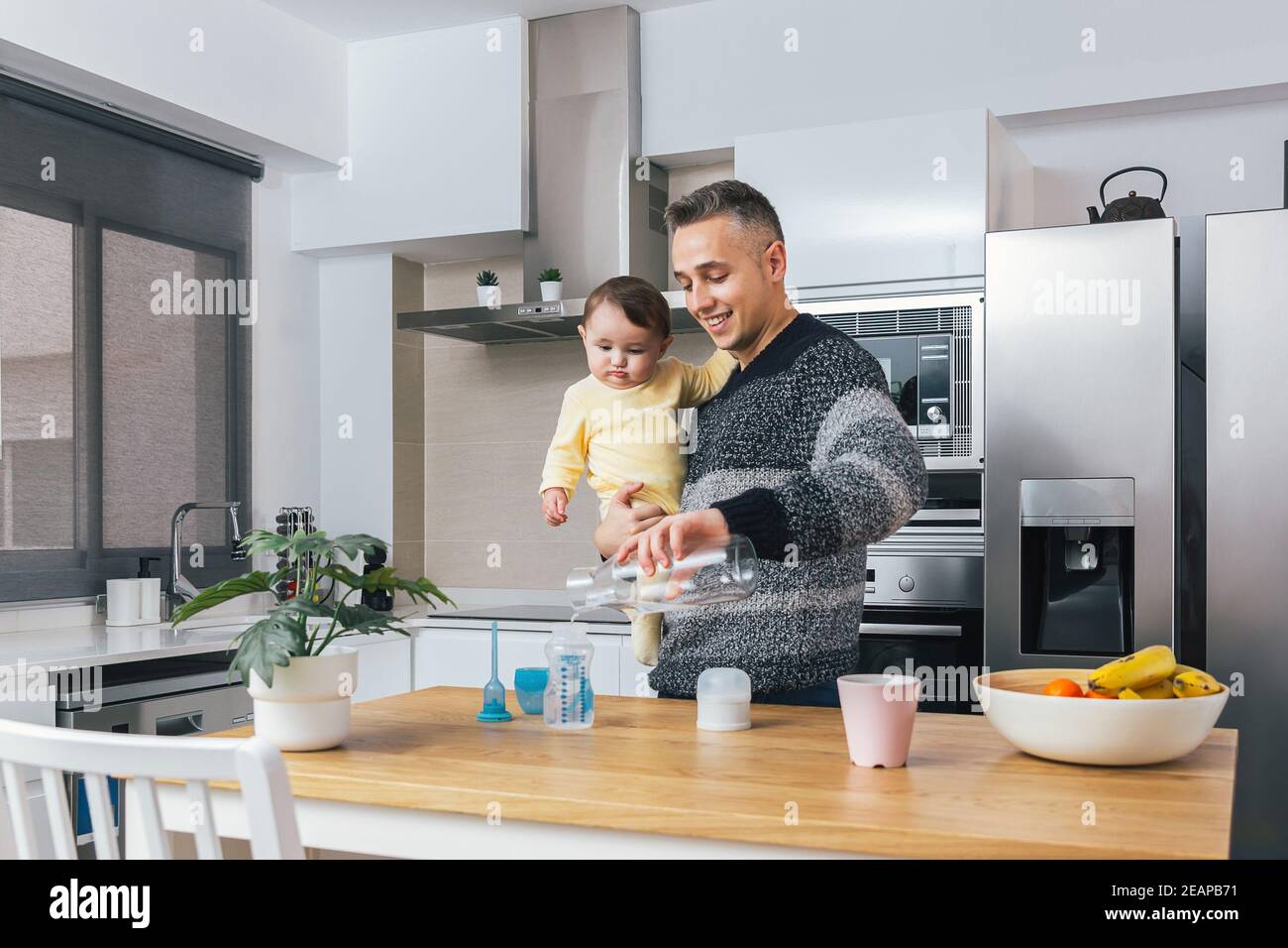 Stock photo of Young single father preparing a feeding bottle while holding his baby in arms in the kitchen. Man and child, parenting fatherhood real Stock Photo