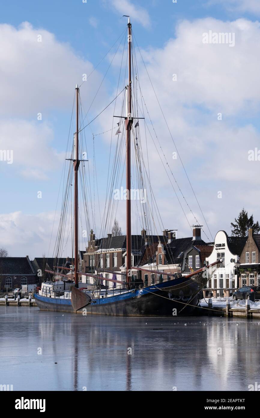 Cityscape of Lemmer with a large sailing yacht with two masts and houses mirrored in the ice of the Dok in the Netherlands in winter Stock Photo