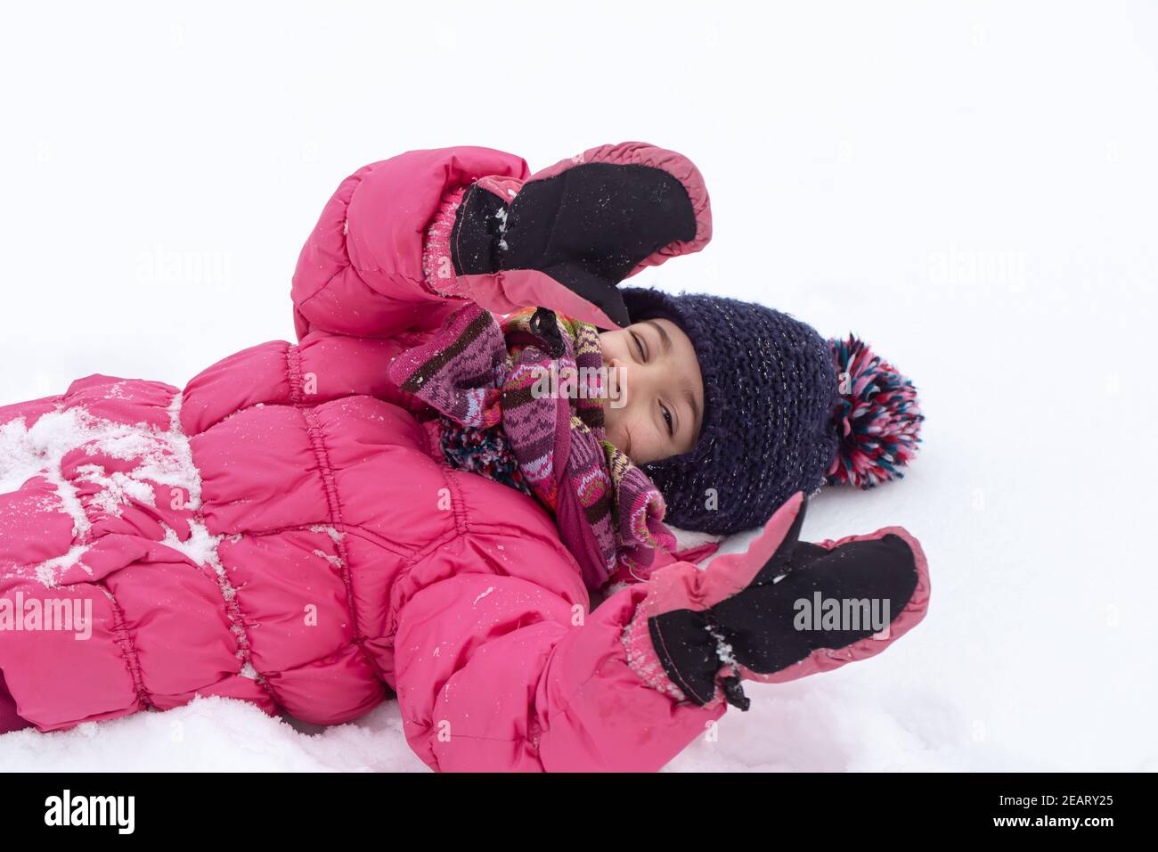 A cute little girl in a pink jacket and a hat is playing in the snow ...