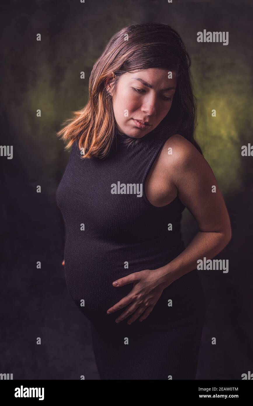 Portrait of a dark-haired pregnant woman in a black tight dress holding her big gestation belly with her hands Stock Photo