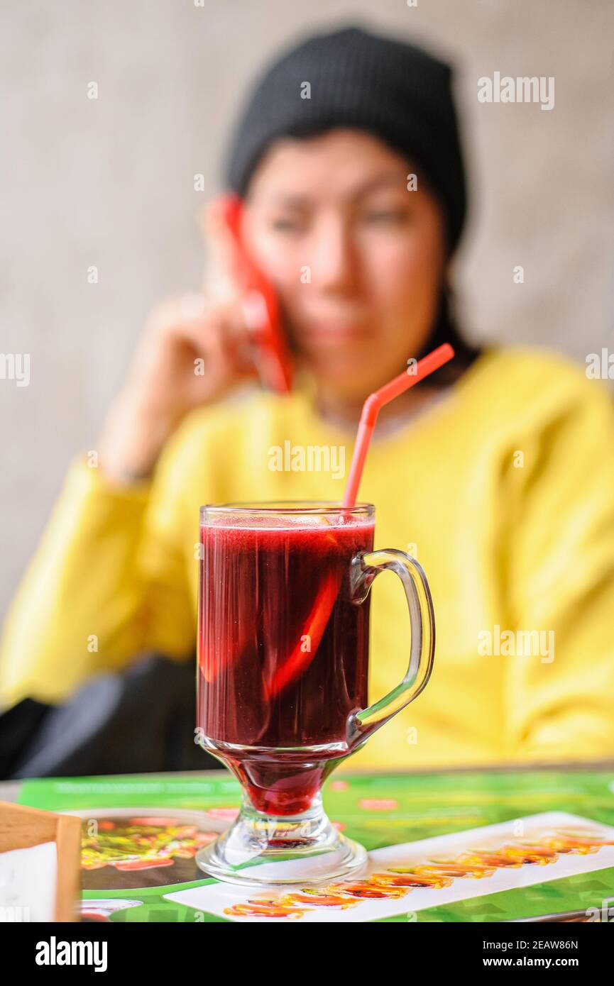 a glass goblet with mulled wine which includes senby red wine  and sliced â€‹â€‹orange with drinking straw stands on a table in a cafe against the background of a young woman who speaks on the phone Stock Photo