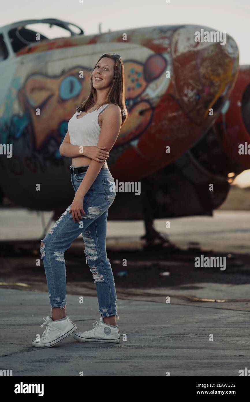 A tall blonde teenage girl wearing  ripped jeans and a white tank top poses for portraits by abandoned airplanes covered in graffiti in the desert wild Stock Photo