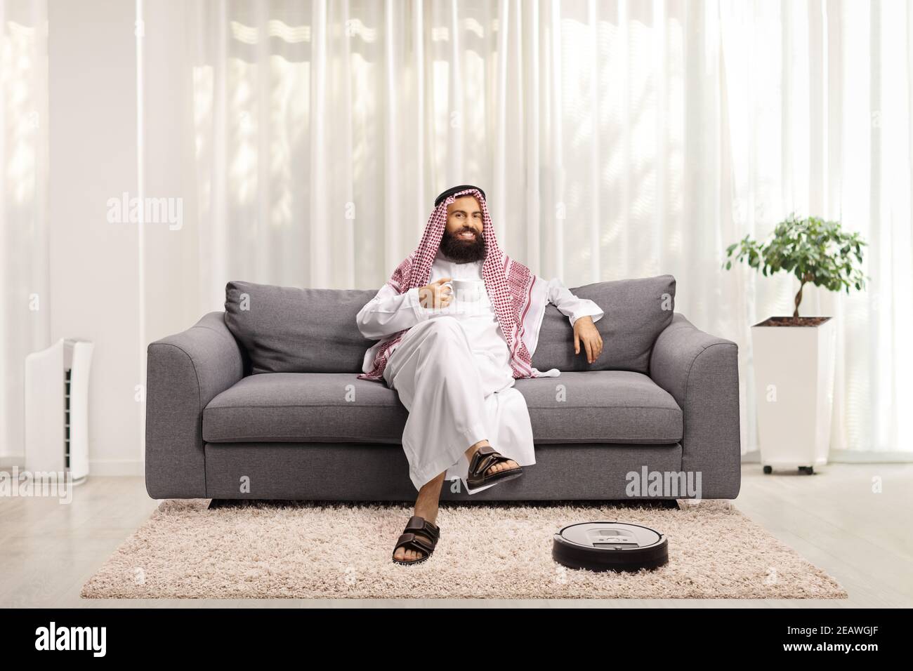 Saudi arab man at home resting on s sofa and a robotic vacuum cleaner