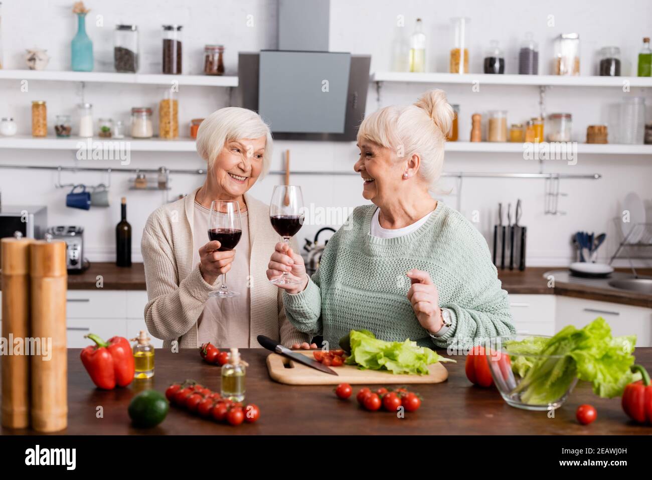 smiling retired women holding glasses of red wine and looking at each other near fresh vegetables on table Stock Photo