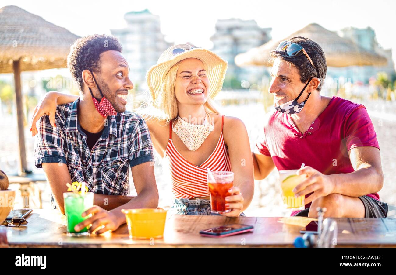 Friends group drinking cocktails at beach with open face masks - New normal vacation concept with people having fun laughing together at chiringuito Stock Photo