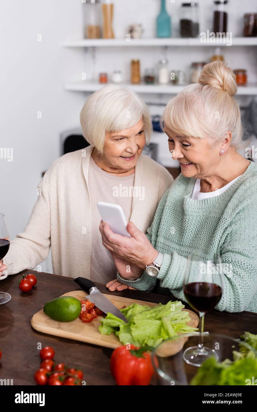 happy senior woman holding smartphone near retired friend in kitchen Stock Photo