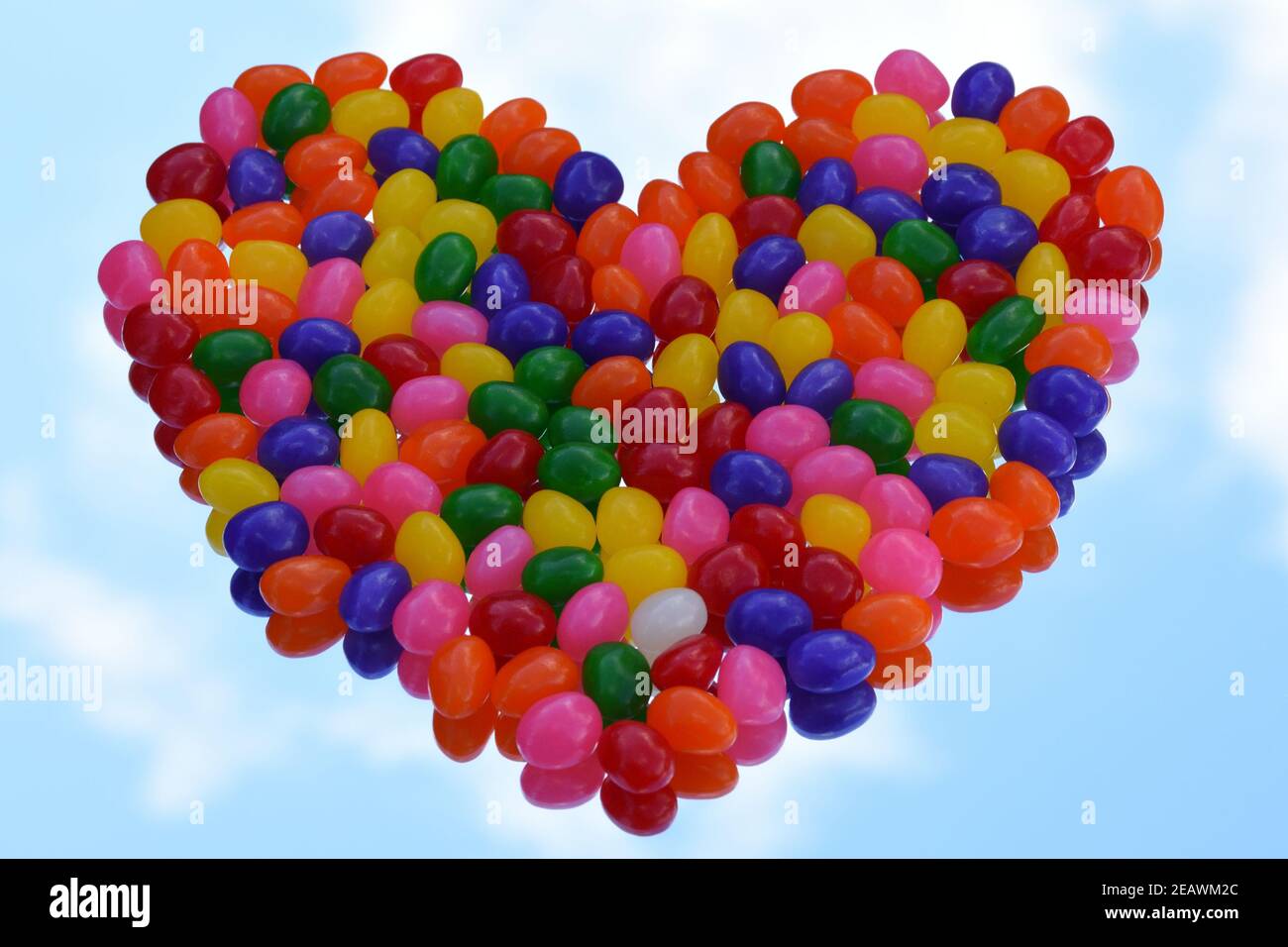 Colorful heart shaped jelly beans on the mirror with the reflection of blue sky with white cloud background. Copy space. 3-dimension. Stock Photo