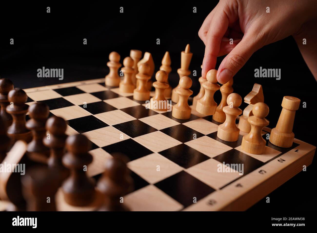 Close up of white and black wooden chess pieces on board. Woman's hand makes first move of white pawn on chessboard. Concept of intelligent, logical a Stock Photo