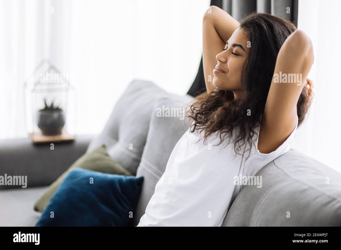 A satisfied young african american woman is relaxing on the couch in living room at home, smiling. Girl takes a break from online distant work, or hav Stock Photo