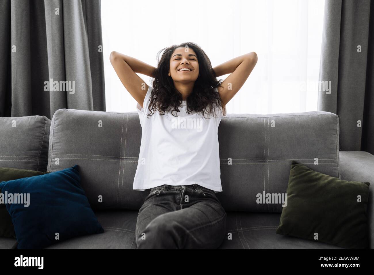 A satisfied young african american woman is relaxing on the couch in living room at home, smiling. Girl takes a break from online distant work, or hav Stock Photo