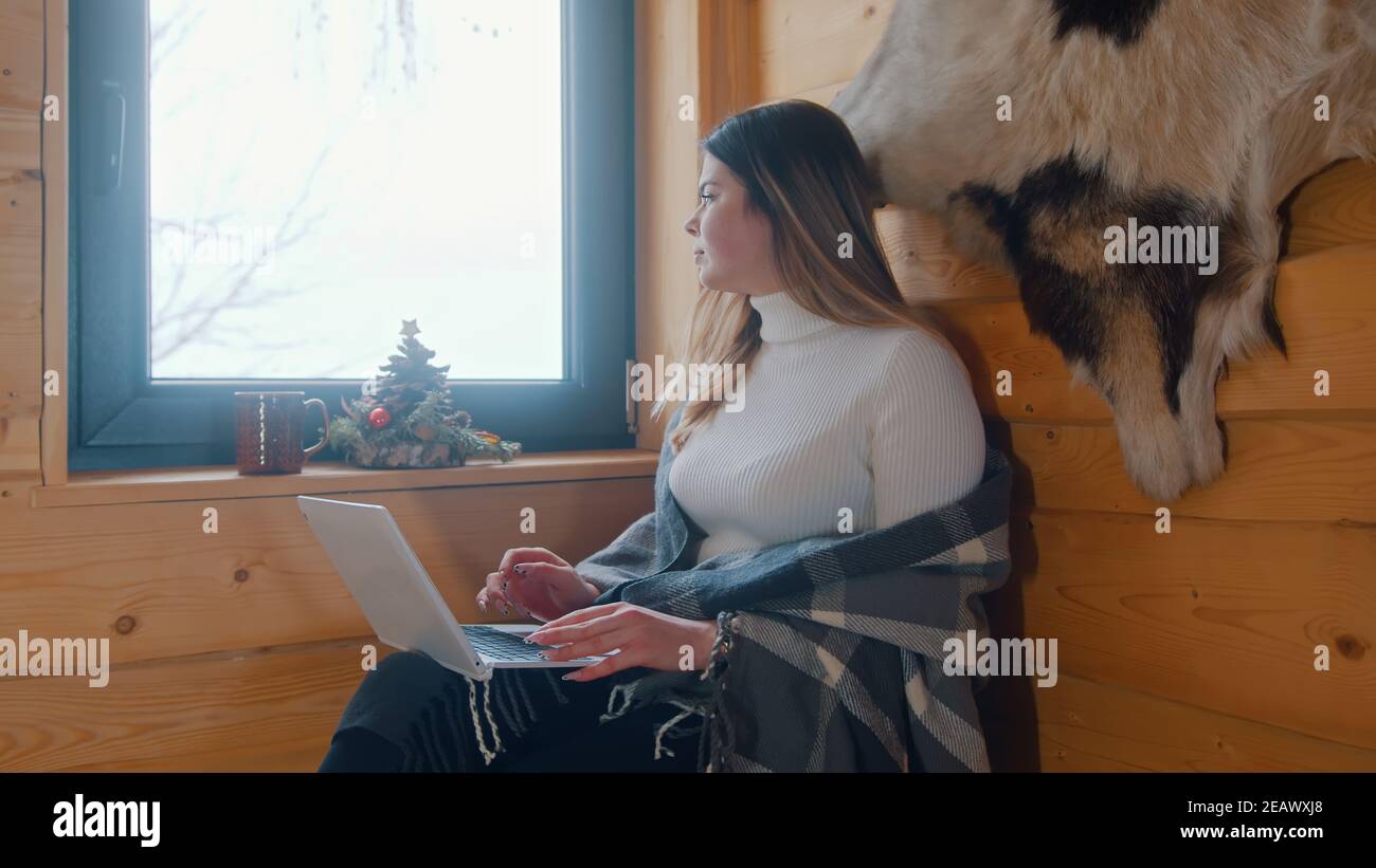 Lonely young caucasian woman sitting near the window covered with blanket lookin outside and using laptop. High quality photo Stock Photo