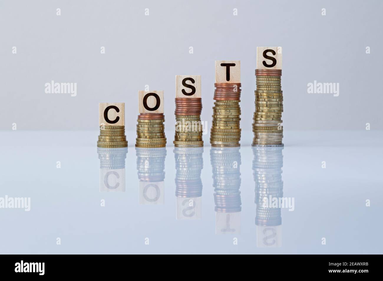 Word 'COSTS' on wooden blocks on top of ascending stacks of coins on gray background. Concept photo of business, cost increase, financial reduction. Stock Photo