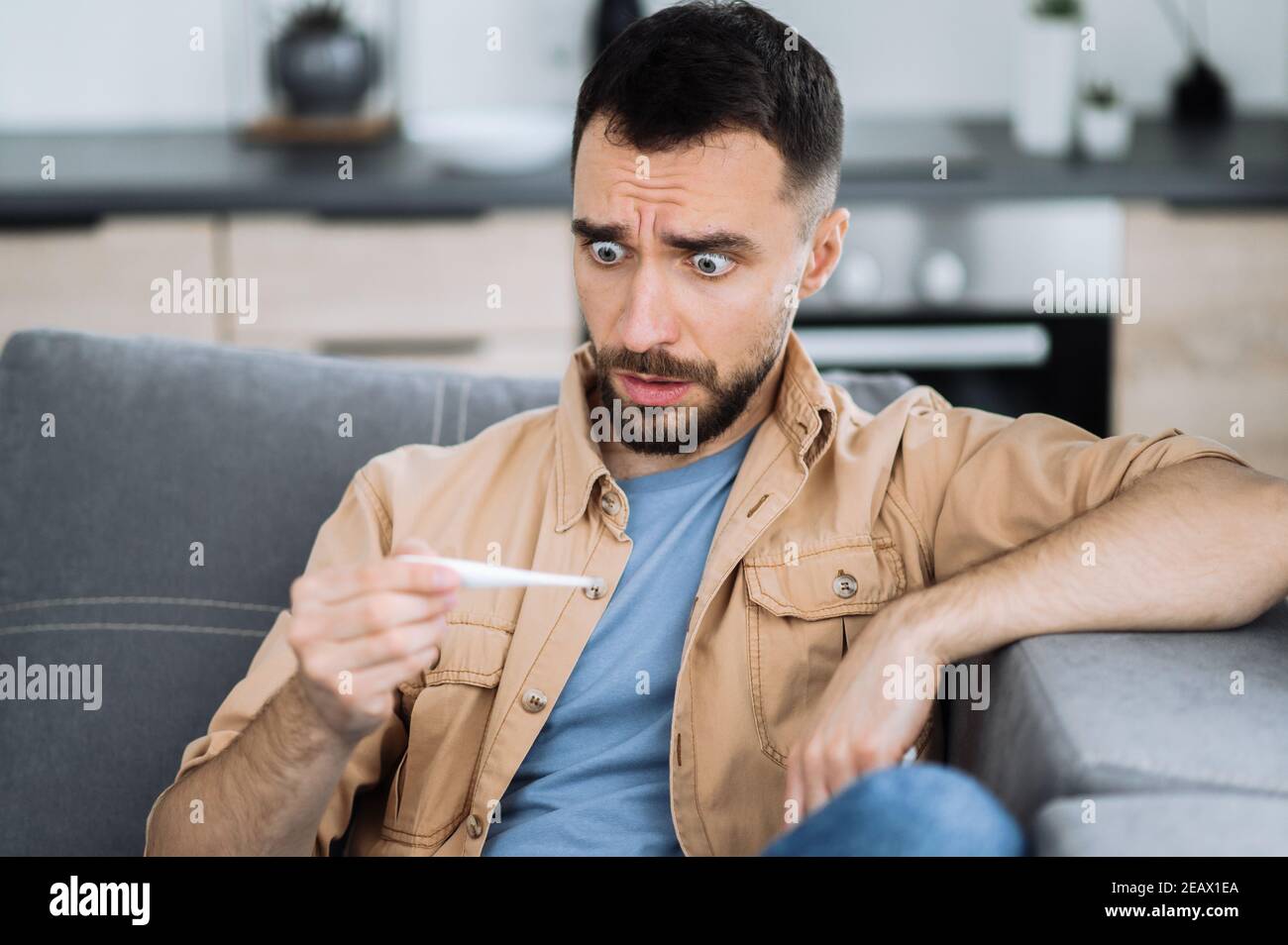 Shocked sick caucasian guy sits on couch feel unhealthy, he frightened looks at the thermometer, ill man with high fever, he has a headache and needs Stock Photo