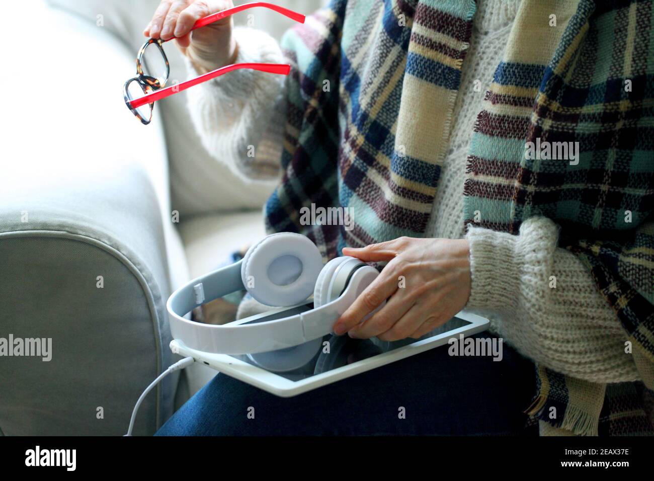 Closeup shot of a woman sitting on the couch holding glasses and headphones with a tablet on her lap Stock Photo