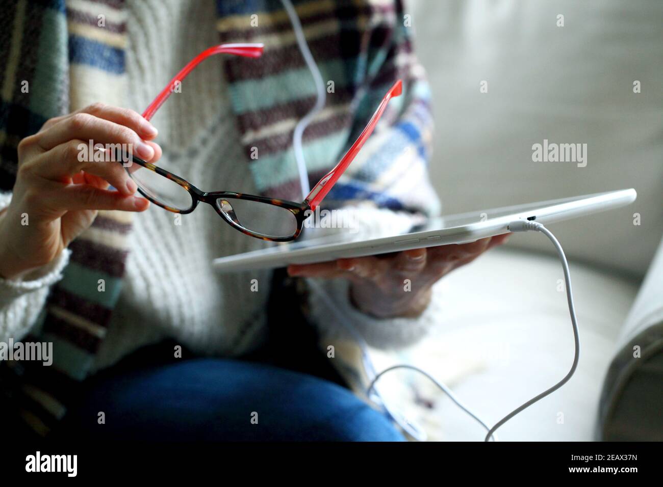 Closeup shot of a woman sitting with headphones on the couch and holding glasses next to a tablet Stock Photo