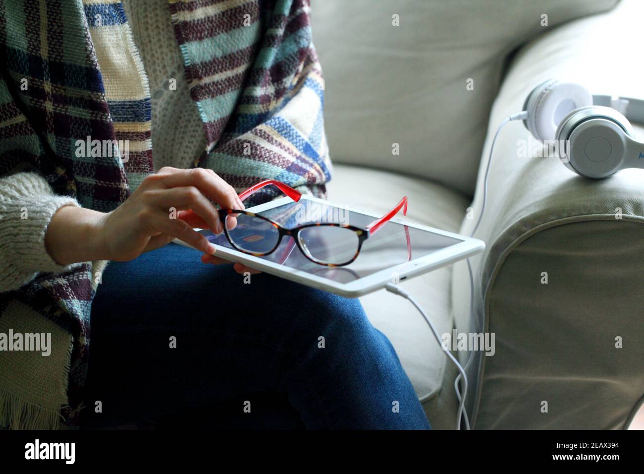 Closeup shot of a woman sitting with headphones on the couch and holding glasses next to a tablet Stock Photo