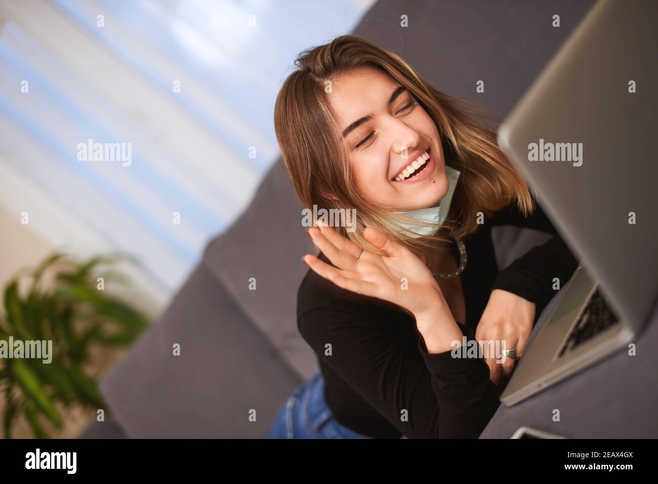 Caucasian white Teenager girl having a video call from computer and a facemask while lying down on a gray couch in house. Stock Photo