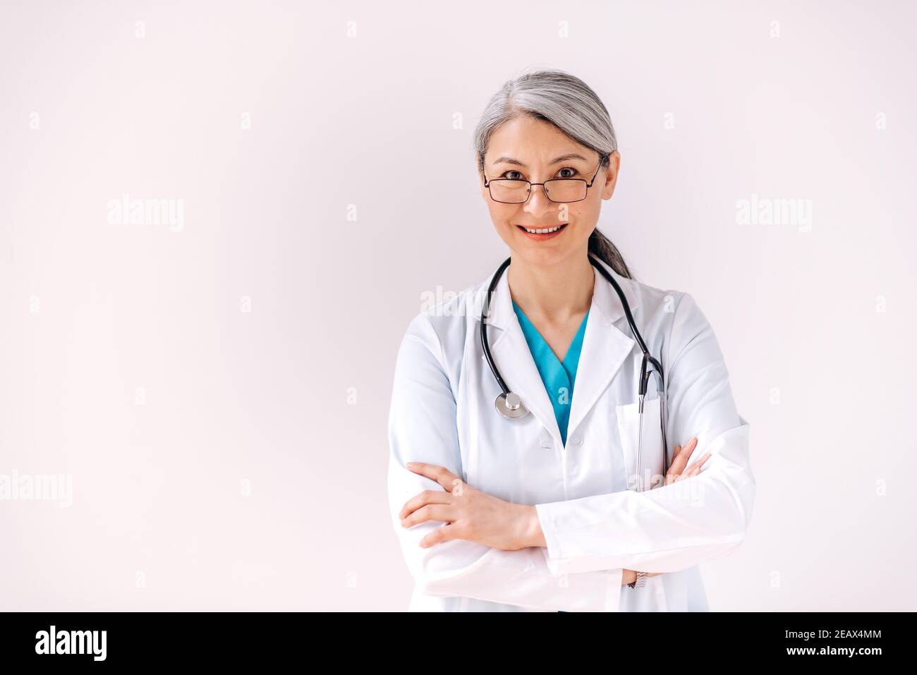 Portrait of a confident asian gray-haired female doctor in medical uniform and stethoscope stands against isolated white background with arms crossed, Stock Photo