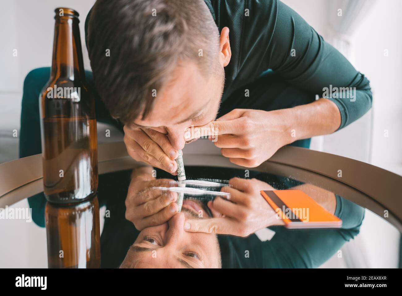 Young addicted junkie man sniffing or snorting cocaine lines on mirror through rolled banknote. Narcotics and addiction concept. Stock Photo