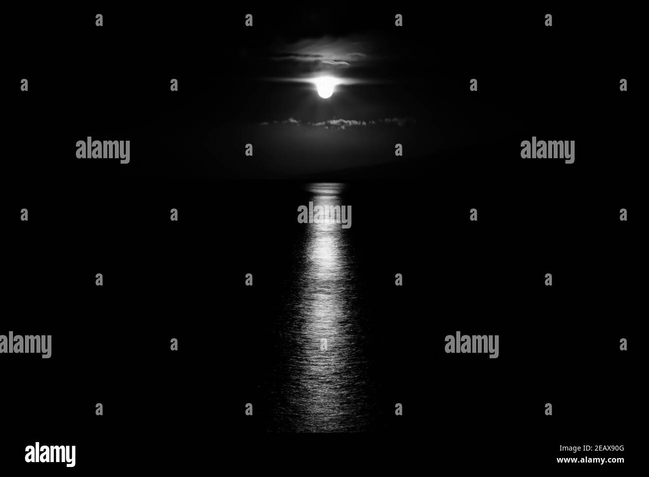 Black and white image of full moon dripping beneath clouds as it sets over the ocean with light reflecting on the sea and moon resembling alien spacec Stock Photo