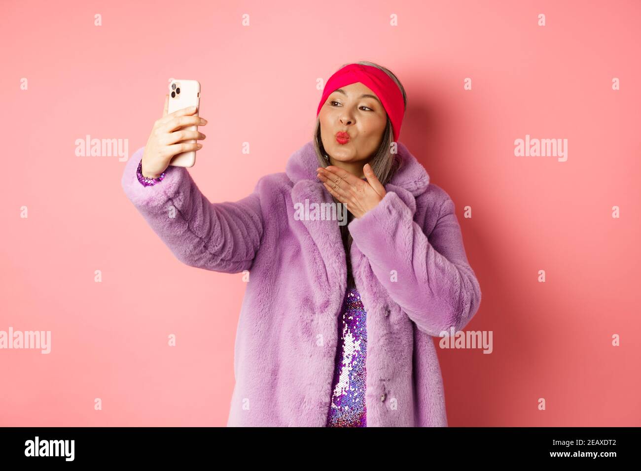 Fashion concept. Beautiful and trendy asian mature woman in faux fur coat with party dress, taking selfie, sending kiss to smartphone camera, posing Stock Photo
