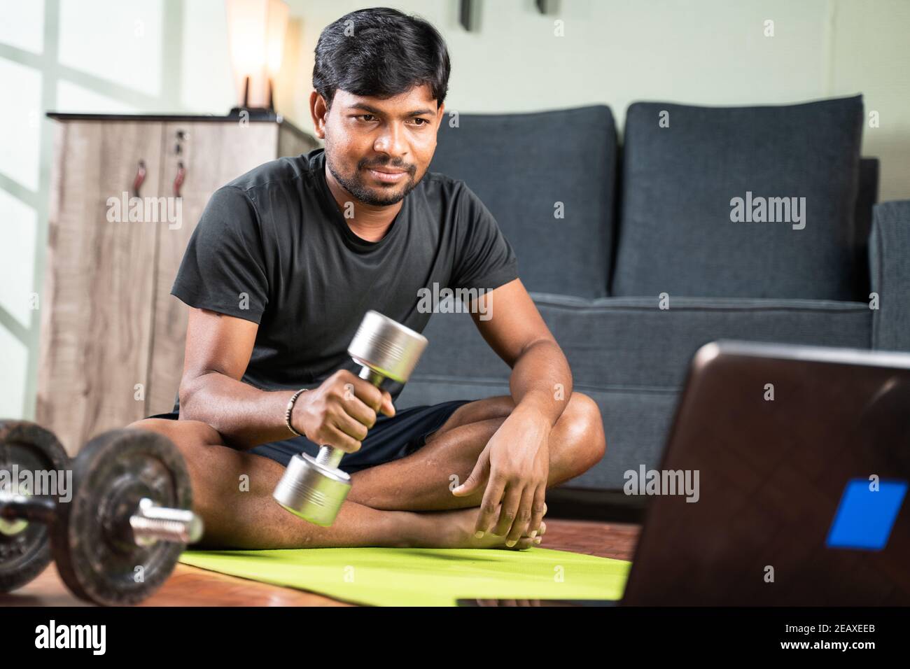 Young man busy learning how to use dumbbells through online tutorials by seeing laptop at home - Concept of Home Gym, exercise due to coronavirus Stock Photo