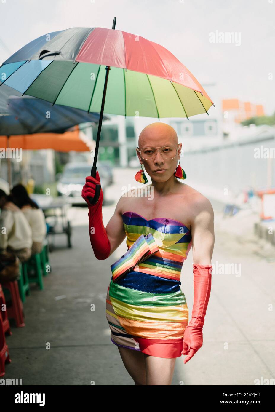 Portrait of an Asian queer person dressed in rainbow colors dress, wearing red gloves and holding a rainbow colors umbrella in Bangkok Thailand. Stock Photo
