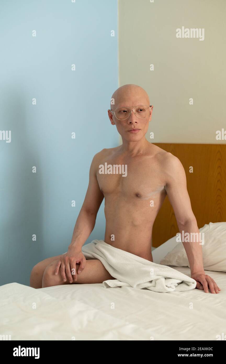 Portrait of a bald Asian man with no clothes except a white towel, sitting  on a bed in a room Stock Photo - Alamy