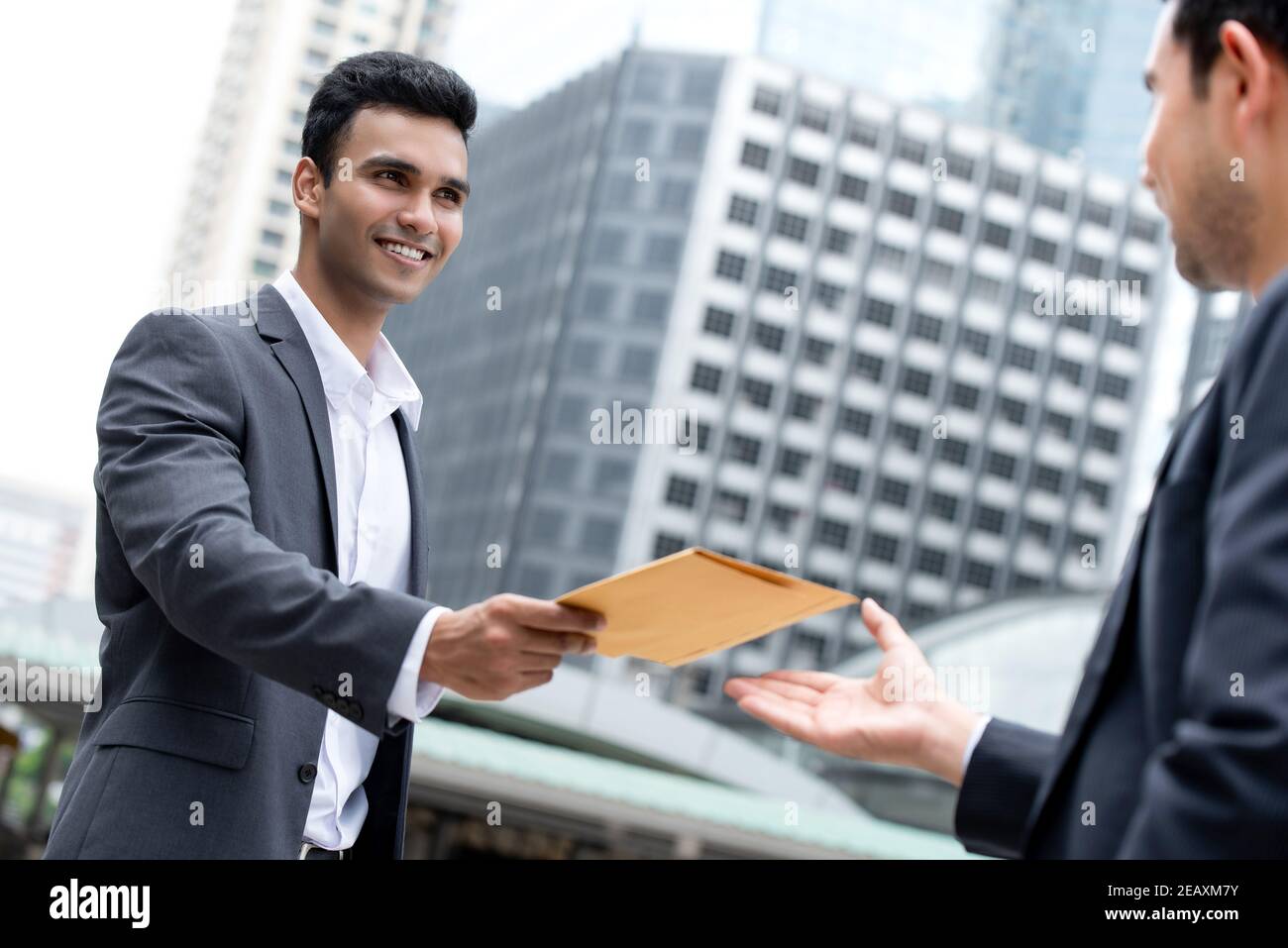 Indian businessman giving document in the envelope to his Asian partner outside office building in the city Stock Photo