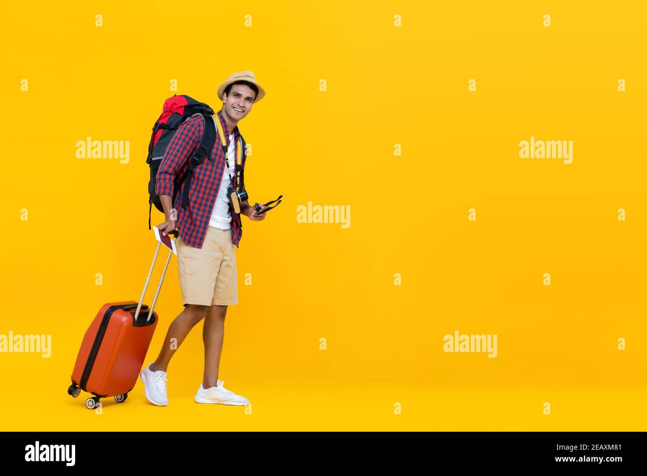 Happy handsome young Asian man with luggage ready to travel for his holidays isolated on yellow studio background with copy space Stock Photo