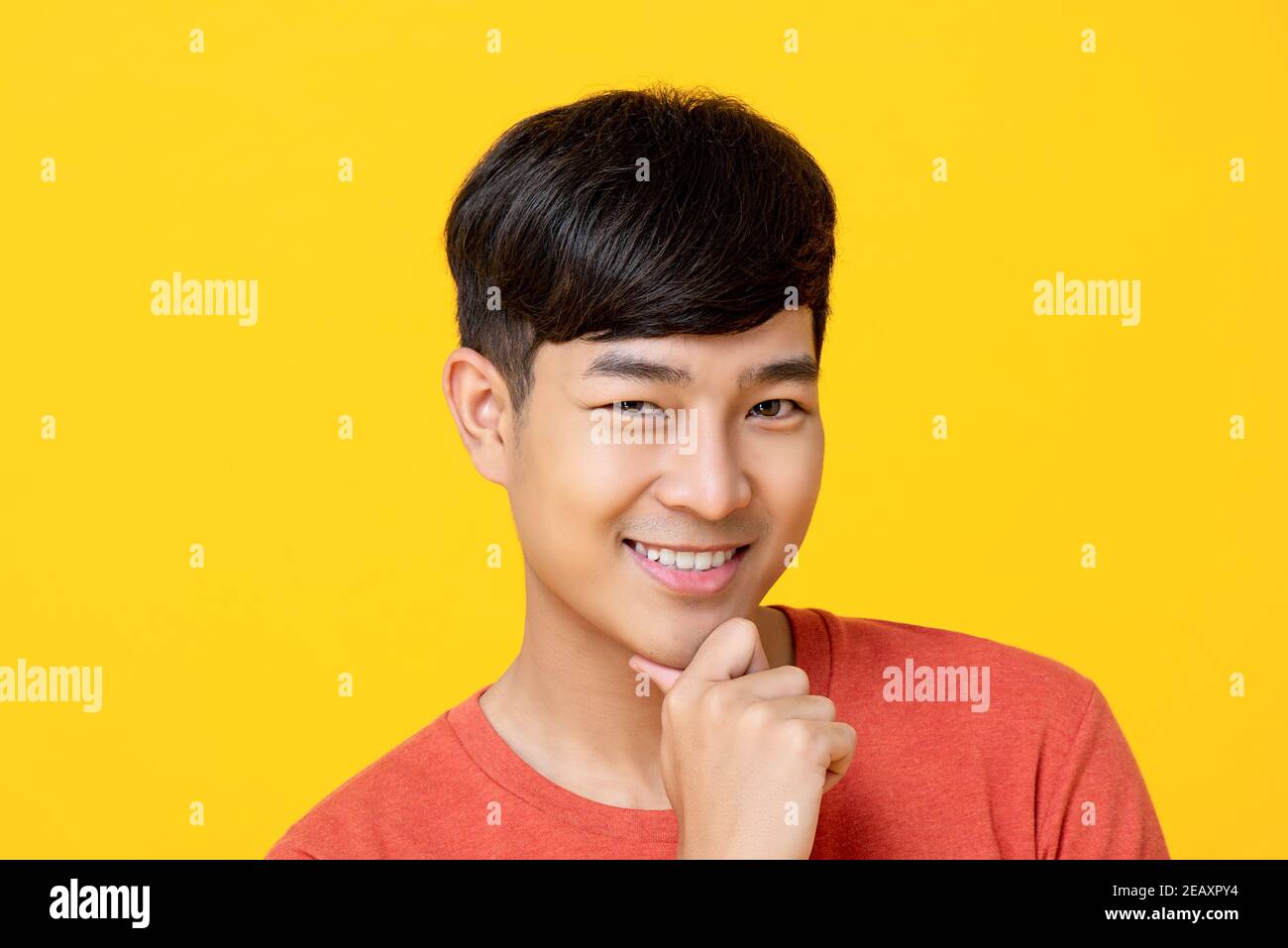 Handsome young Asian man smiling with hand on chin isolated on colorful yellow backkground Stock Photo