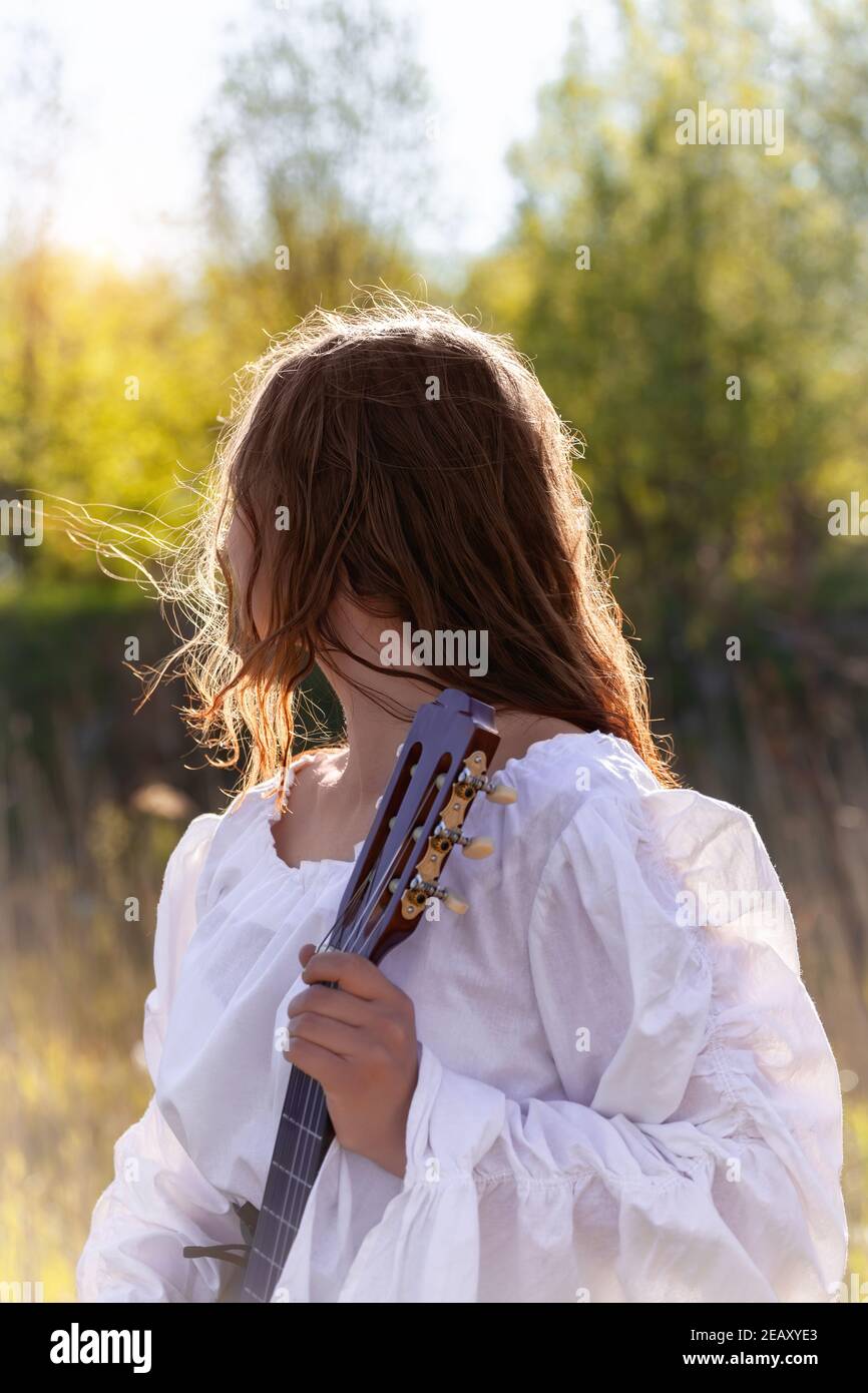 Back view of young woman with red hair in a white medieval dress. Hippie girl holding acoustic guitar at nature. Stock Photo