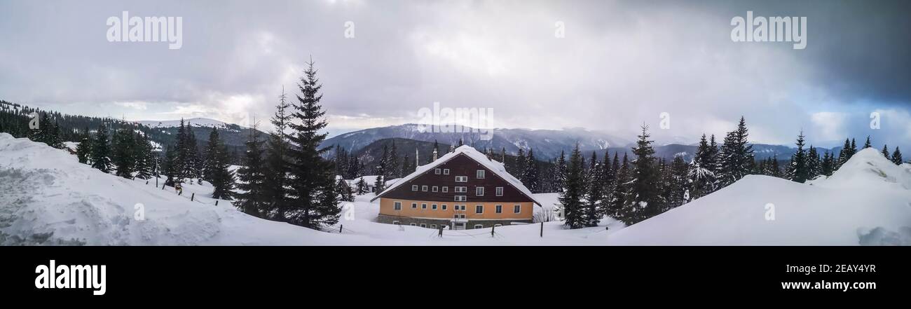 Panorama shot -Winter view overlooking a lodge in the mountains surrounded by evergreens and snow on a cloudy day, in Ranca resort, Romania Stock Photo