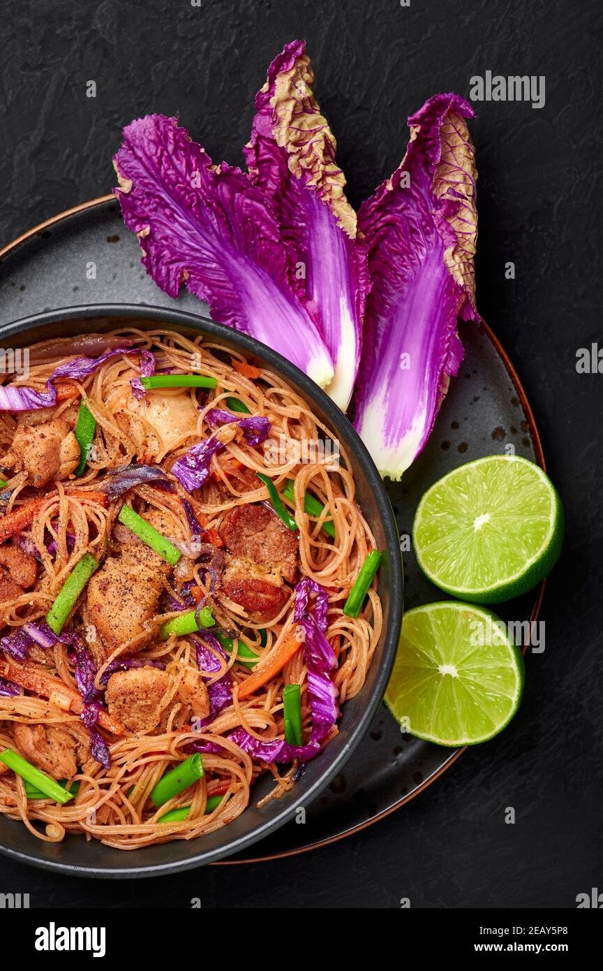 Pancit Bihon in black bowl on dark slate table top. Filipino cuisine noodles dish with pork belly, chicken, vegetables. Asian food. Top view Stock Photo
