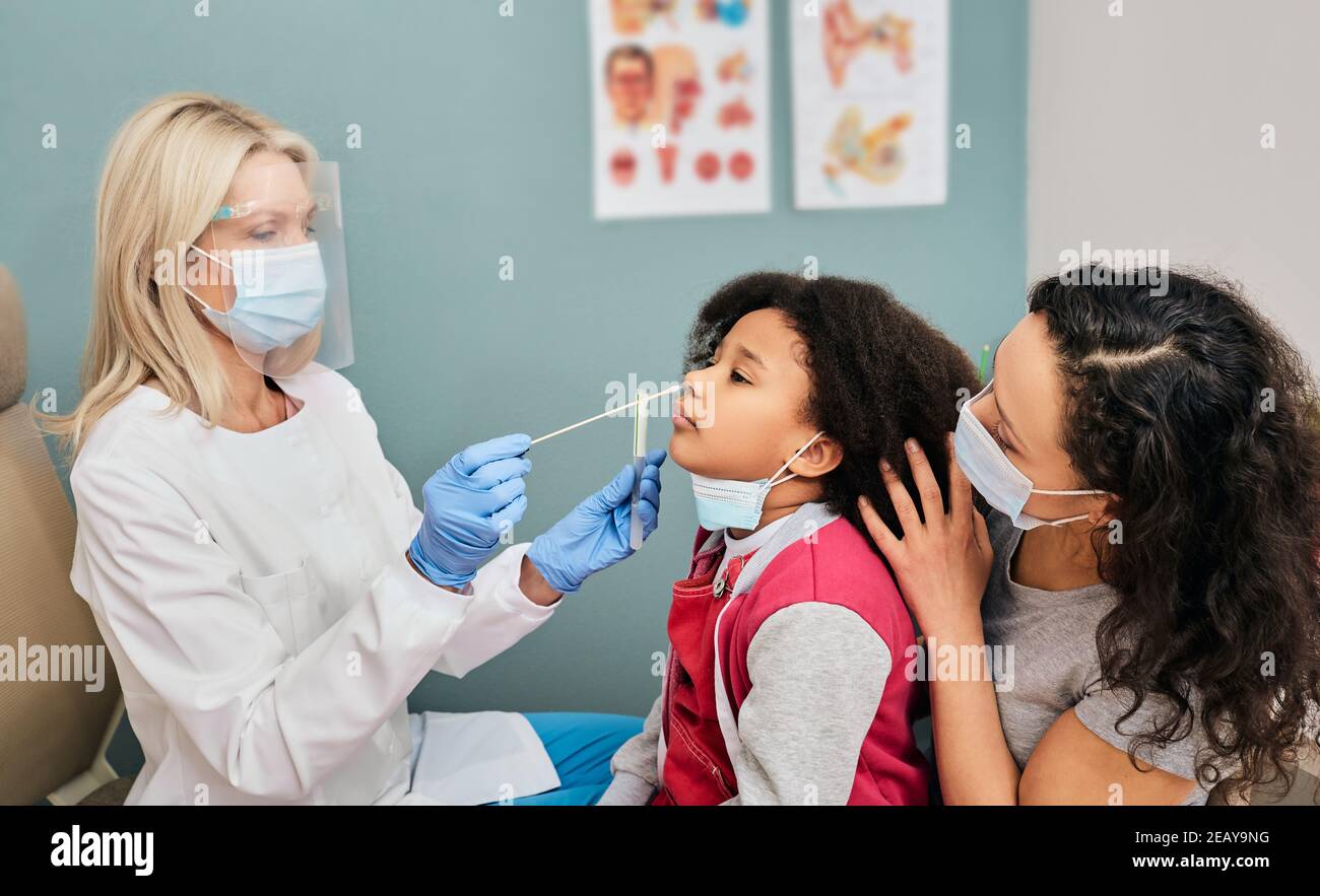 African American female kid having PCR test during coronavirus epidemic. doctor wearing a protective shield using a cotton swab makes a kid's nasal ca Stock Photo
