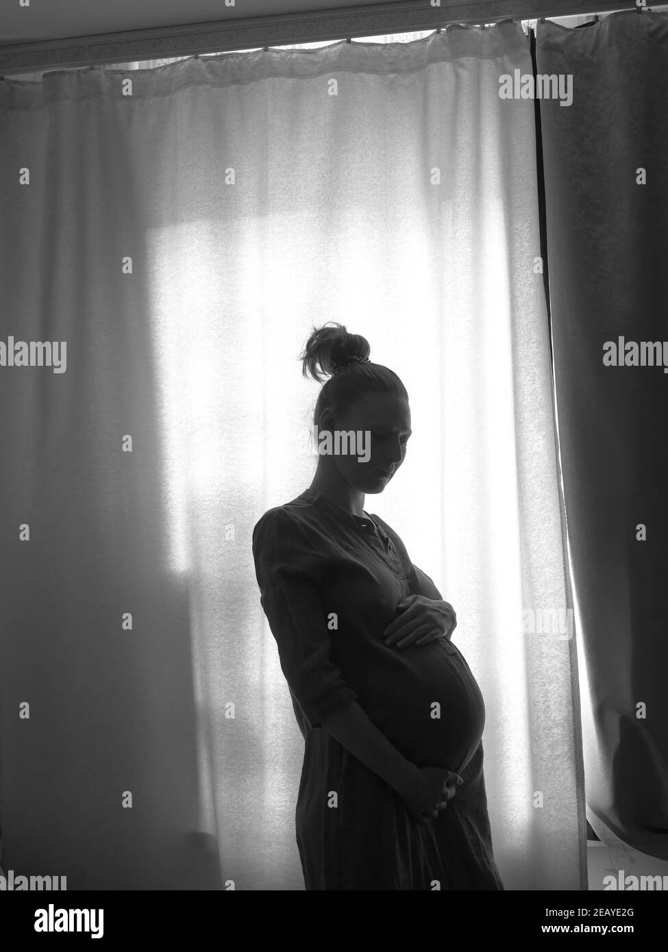 The silhouette of a pregnant girl standing by the window. Black and white photo. Stock Photo