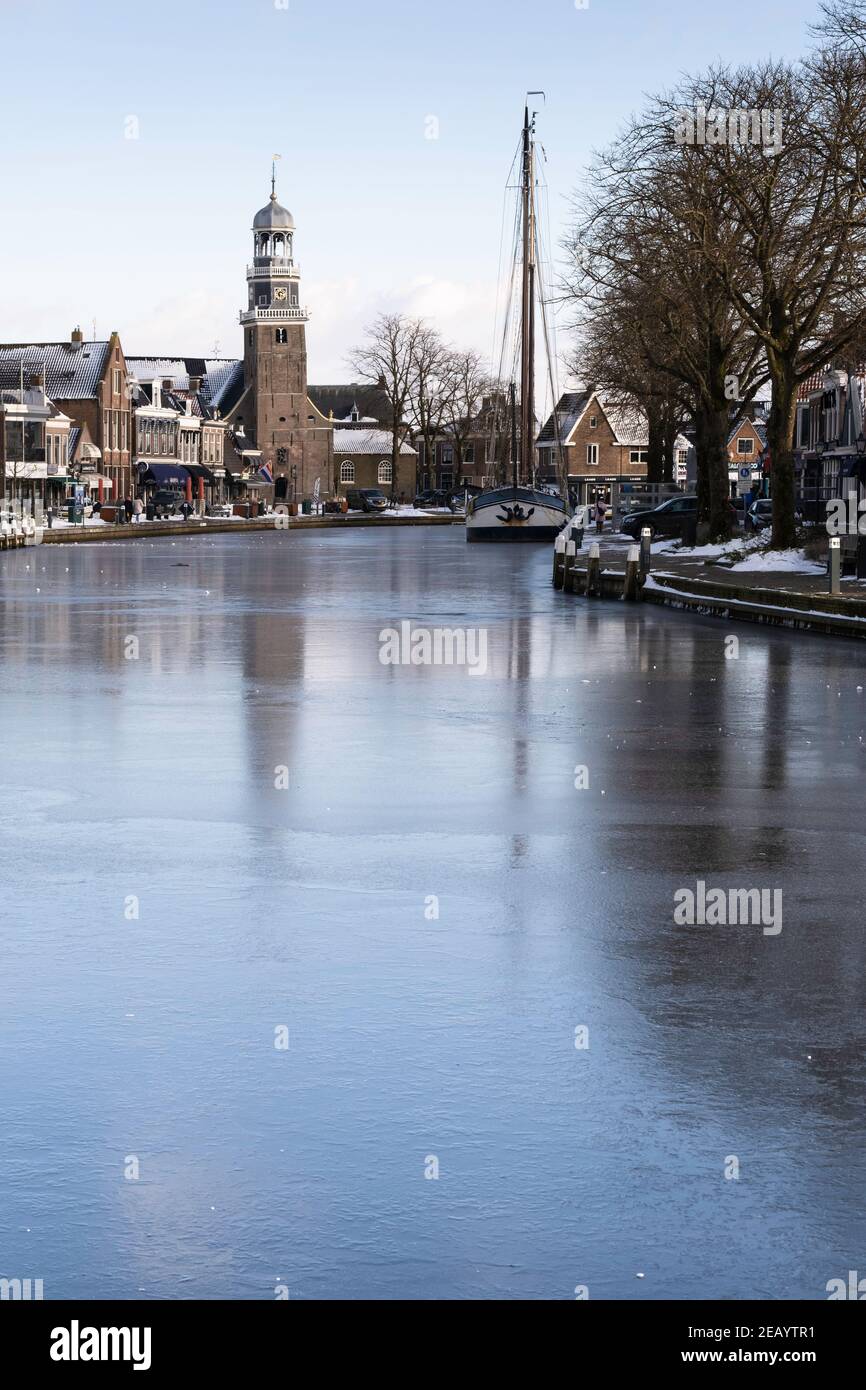 Winter Cityscape of Lemmer, the Netherlands with Reformed Church and shops on the quay with a ship frozen in the ice of Het Dok, blue sky and sun Stock Photo