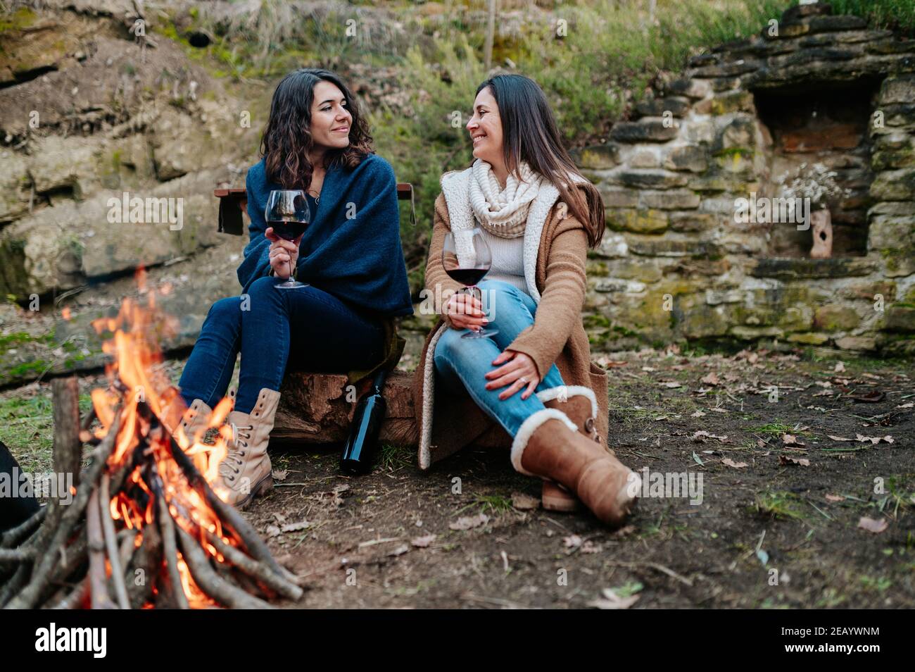 young happy women talking together while drinking glass of red wine. Females warming next to the fire. Campfire, outdoors activities concept. Stock Photo
