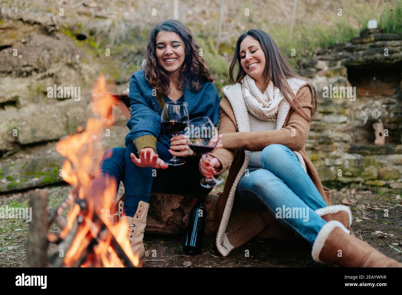 young happy women drinking glass of red wine. Females warming next to the fire. Campfire, outdoors activities concept. Stock Photo