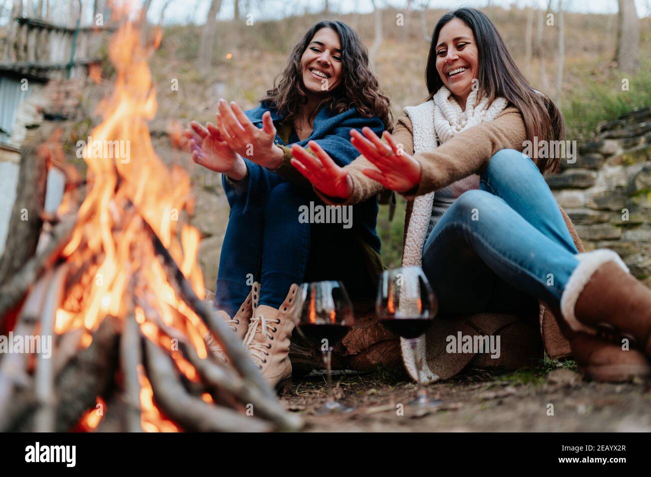 young smiling women warming next to the fire. Glasses of red wine. Campfire, outdoors activities, relaxation, togetherness concept. Stock Photo