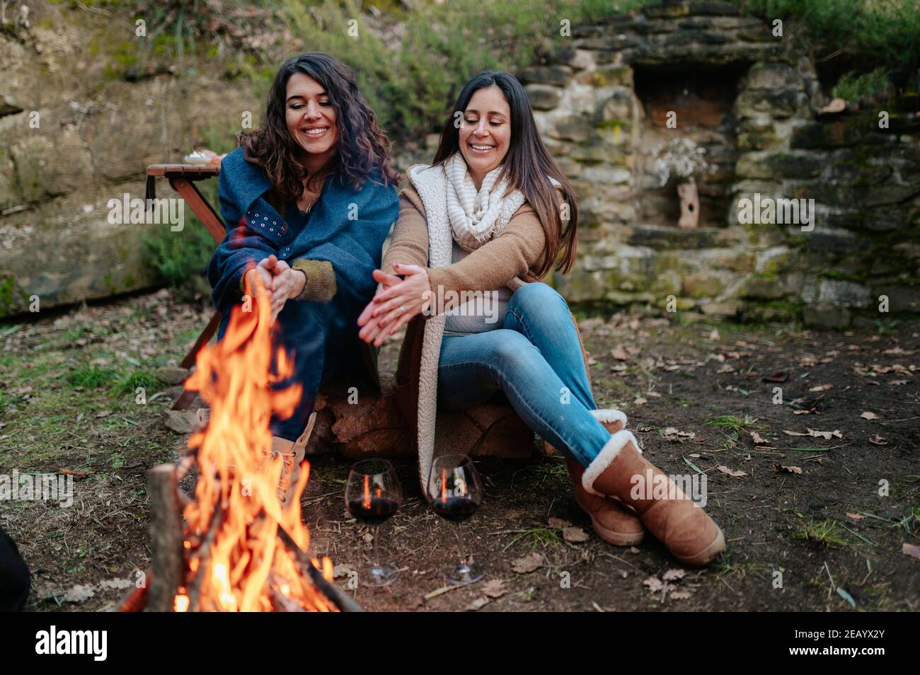 young happy women talking together while drinking glass of red wine. Females warming next to the fire. Campfire, outdoors activities concept. Stock Photo