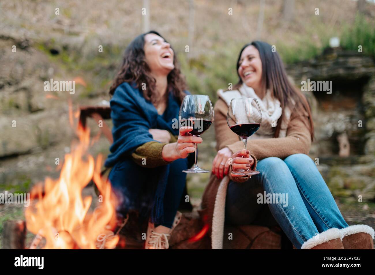 young happy women laughing, holding glass of red wine. Females warming next to the fire. Campfire, outdoors activities concept. Stock Photo