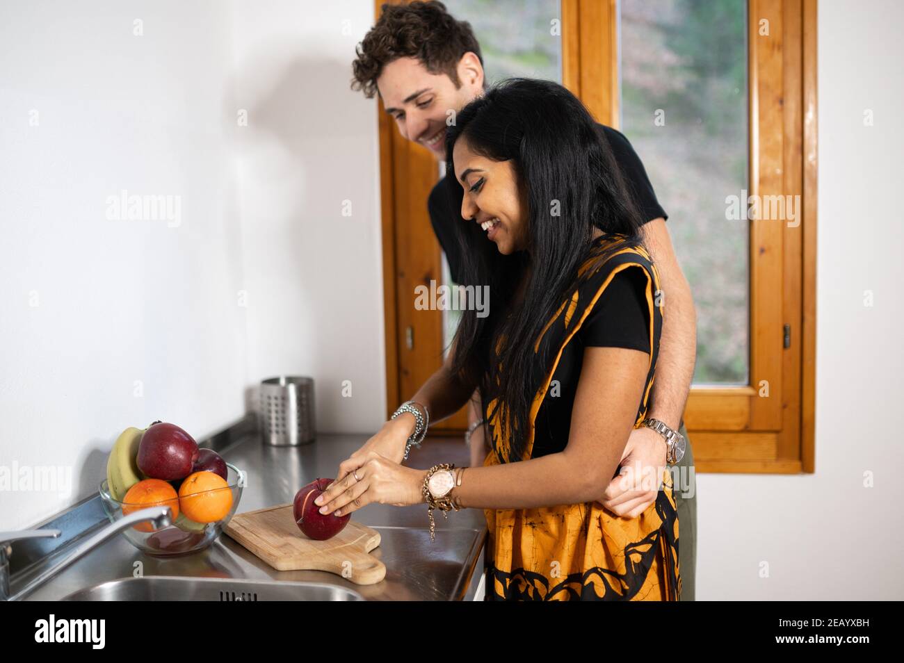 Multi-ethnic couple at home cutting fruit in the kitchen. Indian woman with  traditional dress and caucasian man hugging her from behind Stock Photo -  Alamy