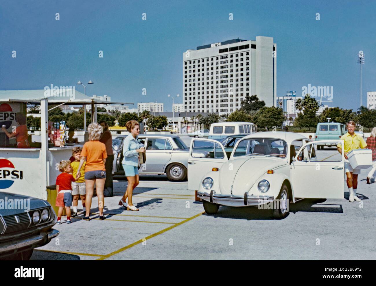 A sunny parking lot in downtown St Petersburg, Florida, USA 1972. A hot day means that it’s shorts weather and good business for the Pepsi Cola kiosk. Opening the car doors of a VW Beetle also helps the interior keep cool. A man carries a polystyrene, insulated cool-box, portable cooler or icebox. At the rear is the Hilton Bayfront hotel. Stock Photo