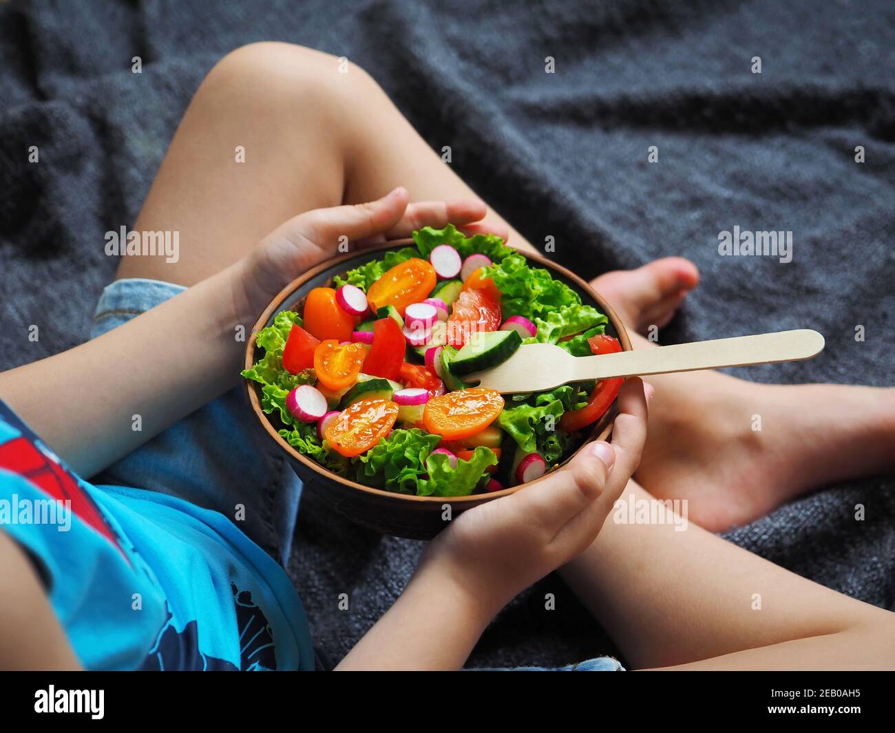 A boy in shorts sits cross-legged on the floor and eats a delicious green salad with orange tomatoes, radish and cucumber. Stock Photo