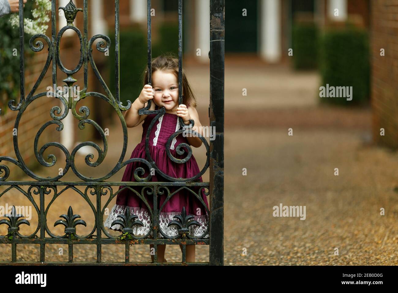 Beautiful two year old girl with a purple dress outside near an iron gate peeking through the gate Stock Photo